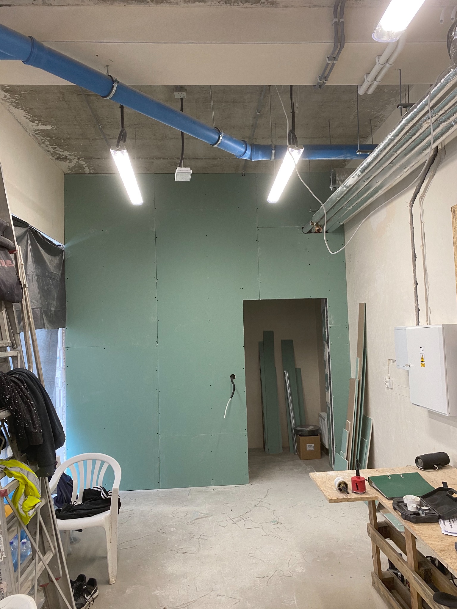 Montaż płyty gipsowo-kartonowej w pomieszczeniu biurowym, widoczne narzędzia na stole roboczym, rury wentylacyjne pod sufitem i oświetlenie budowlane.