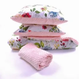 Stos poduszek i zwinięty kocyk z tkaniny w kwiatowy wzór i różowej tkaniny minky, ułożone na białym tle.