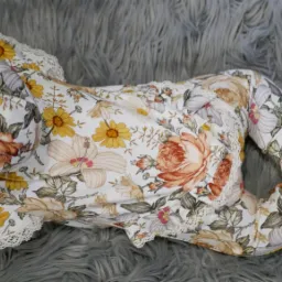 Urocze niemowlę w kwiecistym pajacyku z koronkowym wykończeniem, leżące na puszystym, szarym dywanie.