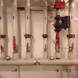 Rozdzielacz instalacji grzewczej z rurami, zaworami, manometrami i pompą obiegową Grundfos, strzałki wskazują kierunek przepływu.