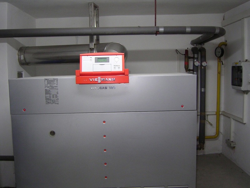 Szary piec gazowy Viessmann Vitogas 100 z czerwonym panelem sterowania w kotłowni, widoczne rury i manometry.