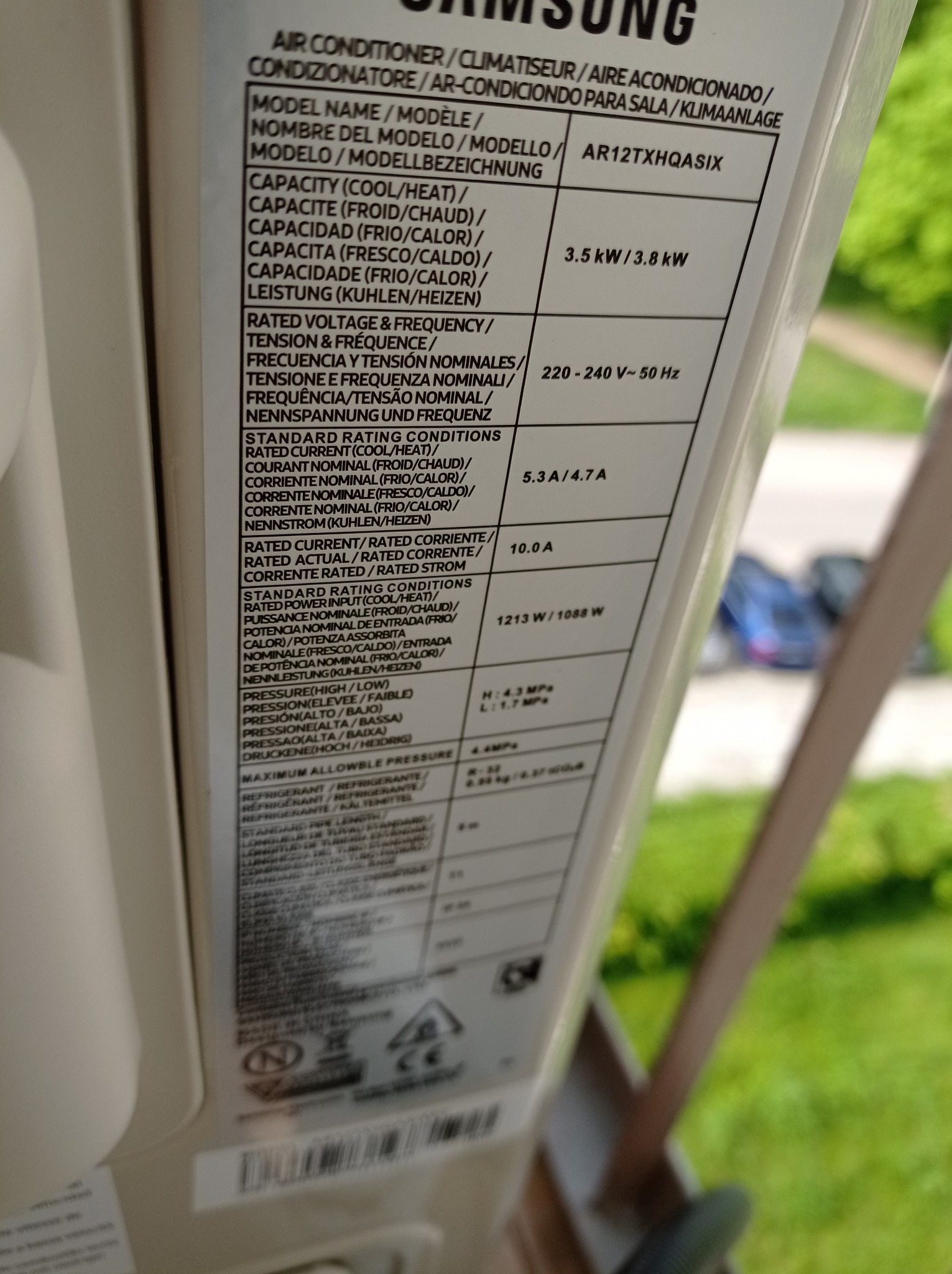 Tabliczka znamionowa klimatyzatora Samsung AR12TXHQASIX z danymi technicznymi w kilku językach, widok z bliska, rozmyte tło zieleni.