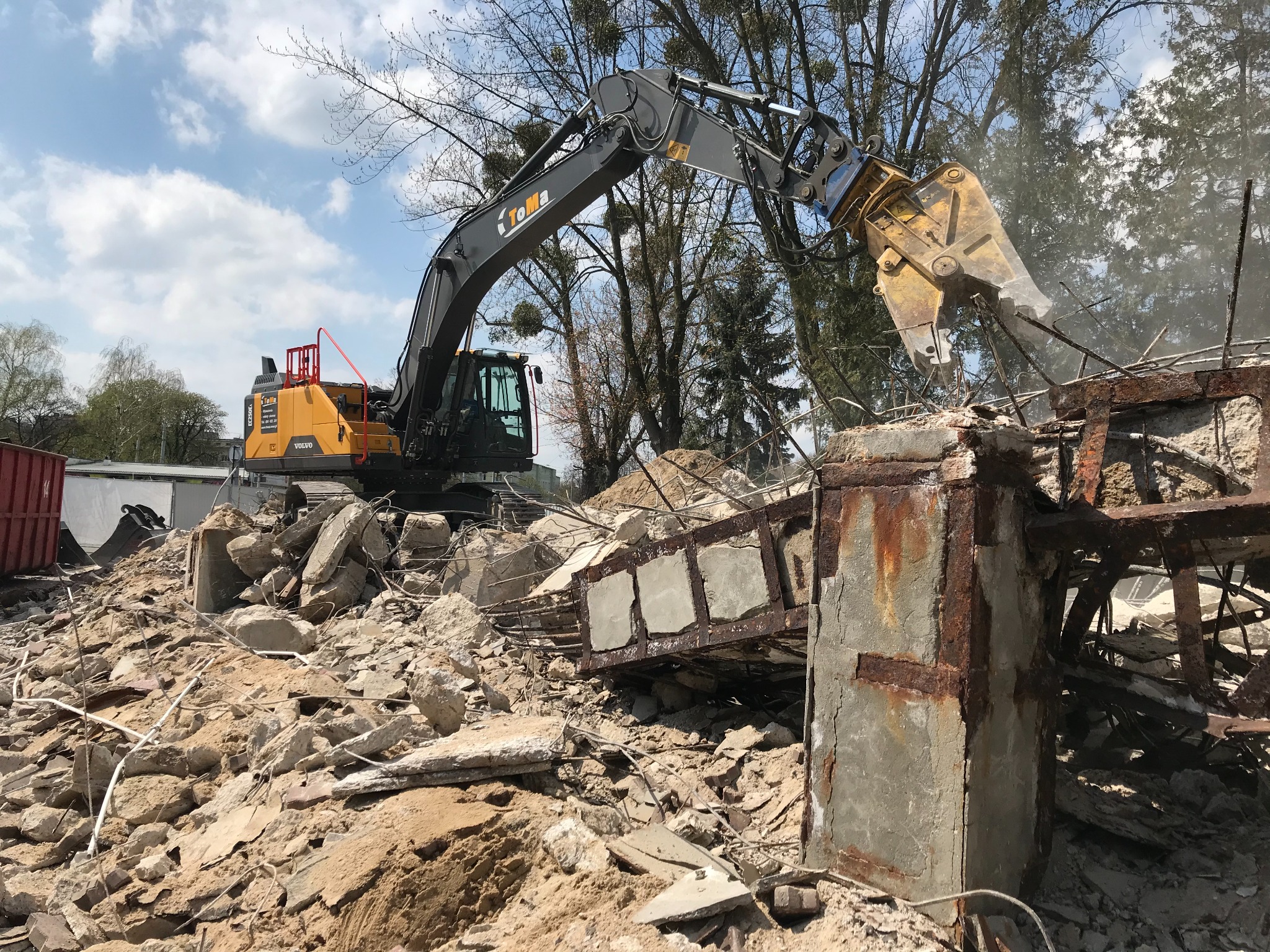 Żółty koparko-ładowarka Volvo z hydraulicznymi nożycami do betonu rozbiera zniszczony, dwupoziomowy podjazd z betonu i stali, z widoczną rdzą i gruzem, na tle drzew i błękitnego nieba z chmurami.