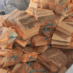 Stos worków siatkowych wypełnionych pociętym drewnem kominkowym, z niebieskimi sznurkami wiążącymi, ułożony na białej plandece, z widocznym zielonym polem w tle.