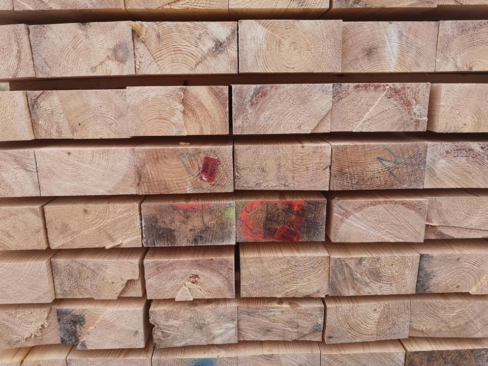 Stos równo ułożonych, przyciętych belek z jasnego drewna z widocznymi słojami, niektóre z czerwonymi oznaczeniami farbą.