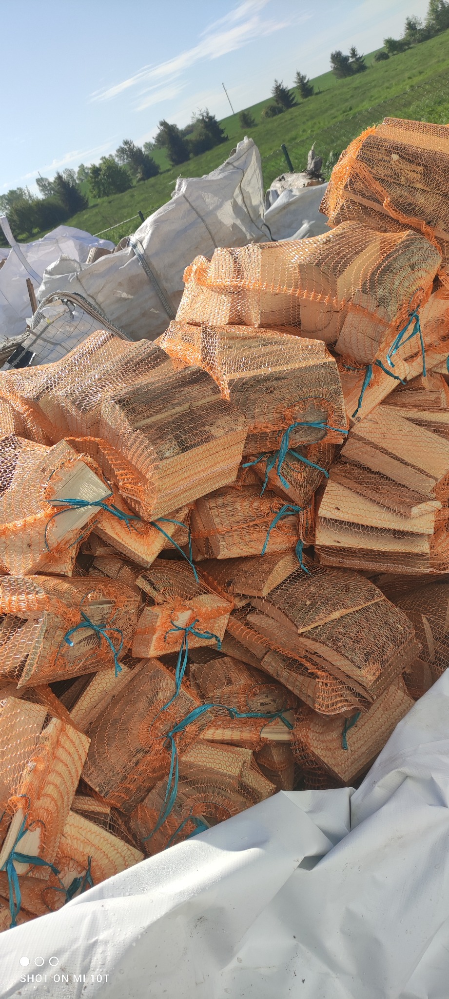 Stos worków siatkowych wypełnionych pociętym drewnem kominkowym, z niebieskimi sznurkami wiążącymi, ułożony na białej plandece, z widocznym zielonym polem w tle.