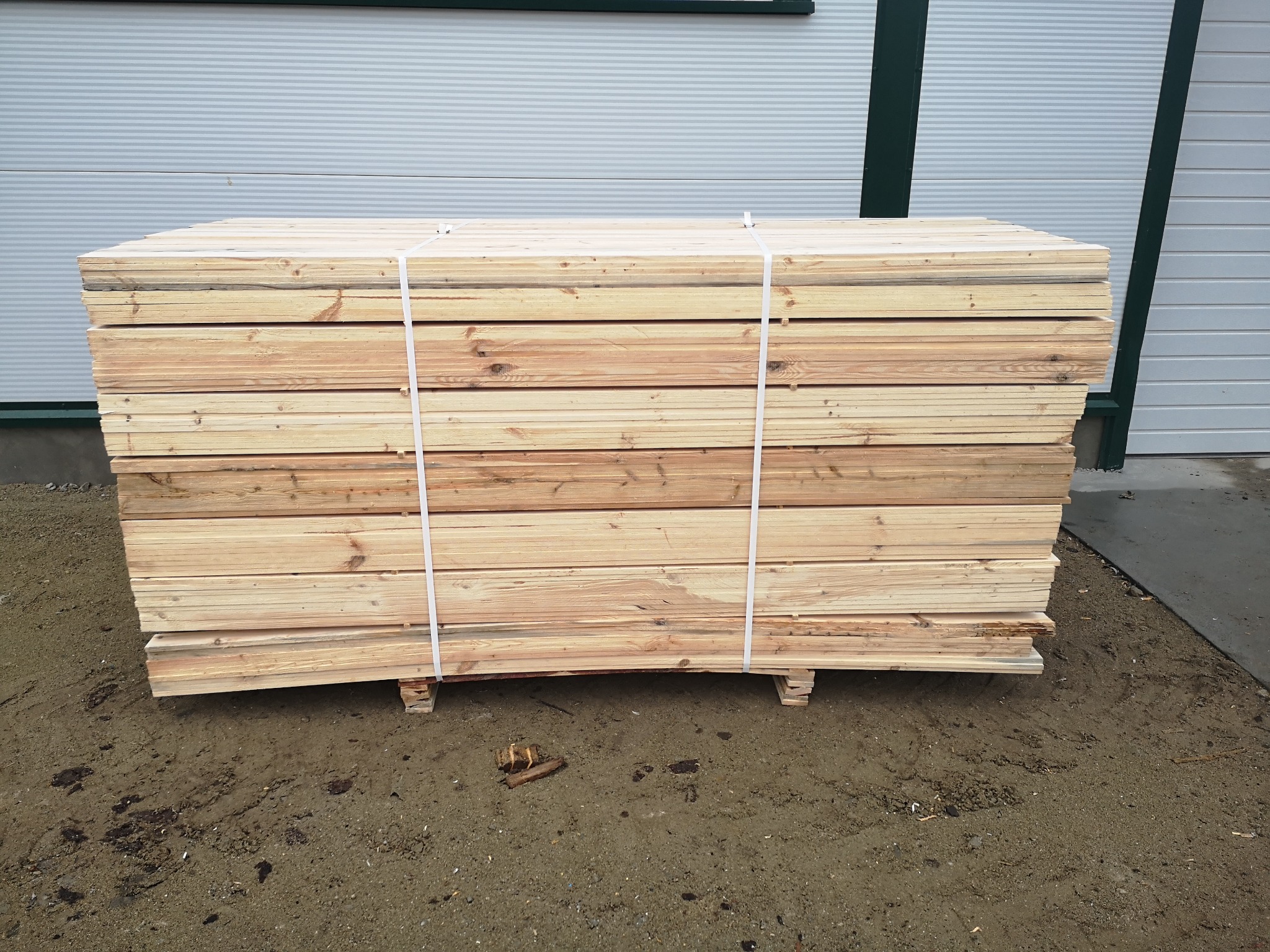 Świeżo oheblowane deski sosnowe ułożone w pakiet i zabezpieczone taśmą, na podkładach, na zewnątrz hali.