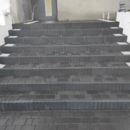 Schody zewnętrzne wyłożone ciemną kostką brukową z jasnymi akcentami na stopniach, prowadzące do drzwi wejściowych.