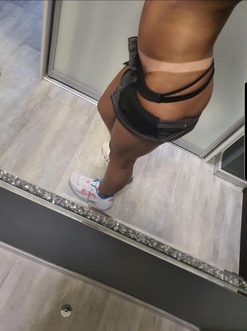 Opalenizna natryskowa na kobiecym ciele, widoczna wyraźna granica opalenizny, ciemne stringi, dżinsowe szorty i białe sportowe buty Fila, odbicie w lustrze z dekoracyjną ramą.