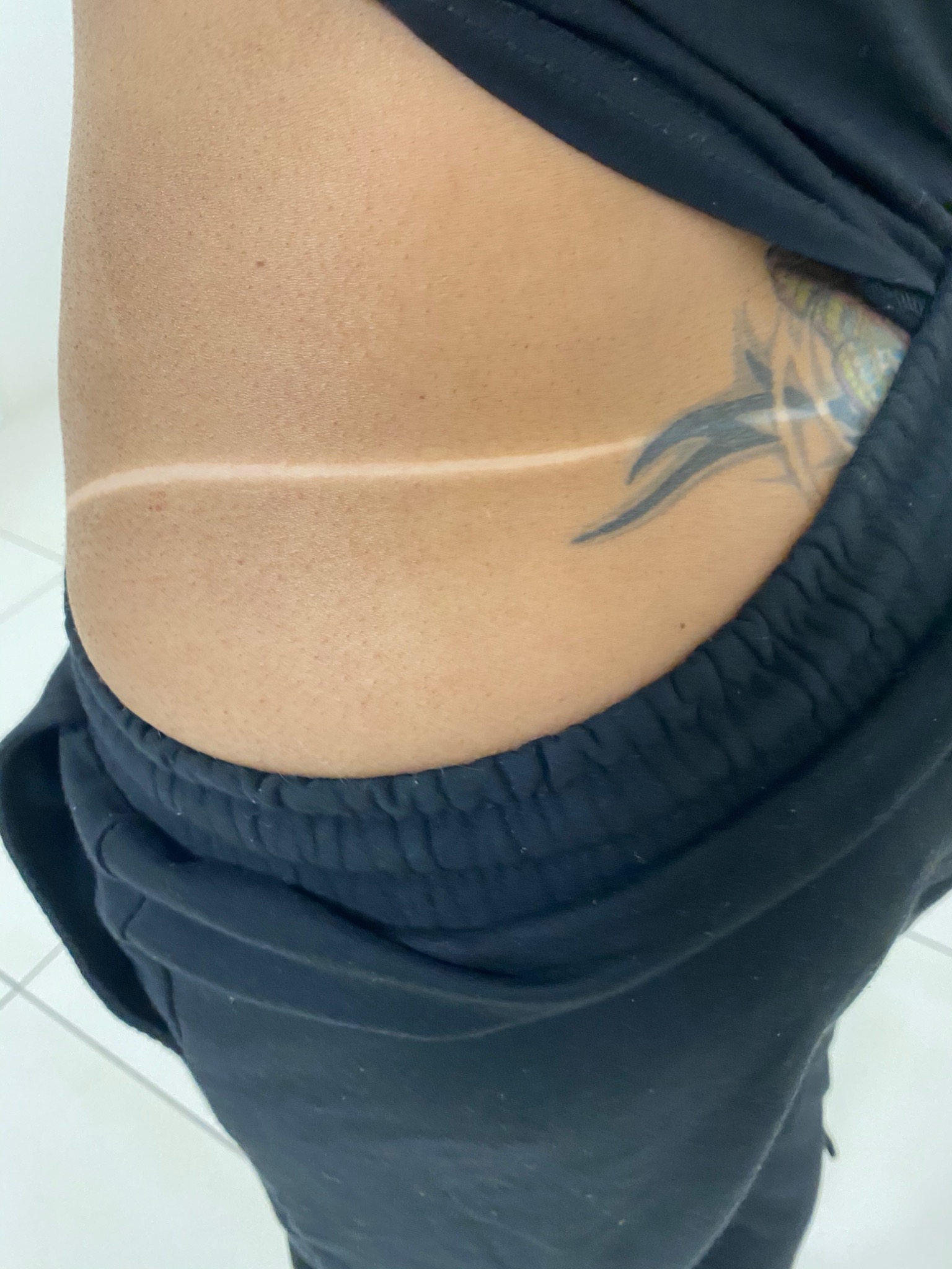 Świeża opalenizna z wyraźną linią, widoczny tatuaż na boku, czarne spodnie dresowe.