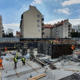 Budowa fundamentów budynku wielorodzinnego z widocznymi zbrojeniami, szalunkami i pracownikami w strojach roboczych, w tle zabudowa miejska i żuraw.