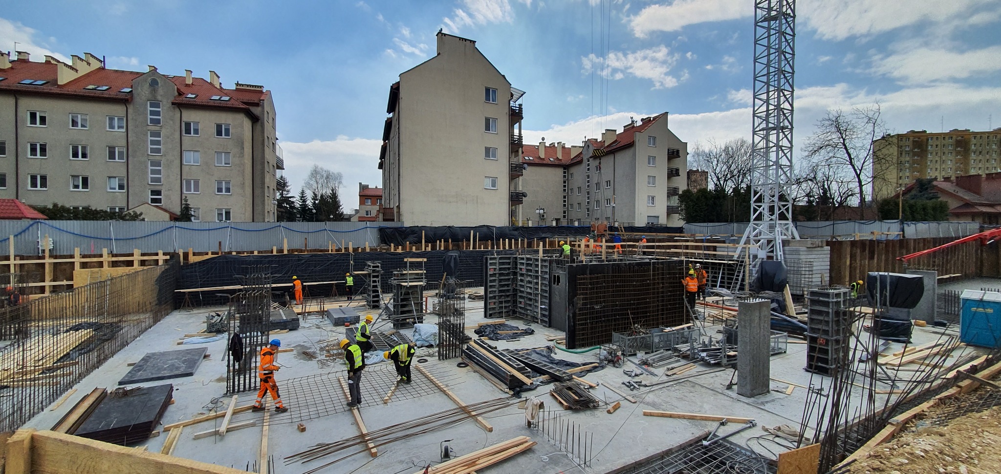 Budowa fundamentów budynku wielorodzinnego z widocznymi zbrojeniami, szalunkami i pracownikami w strojach roboczych, w tle zabudowa miejska i żuraw.