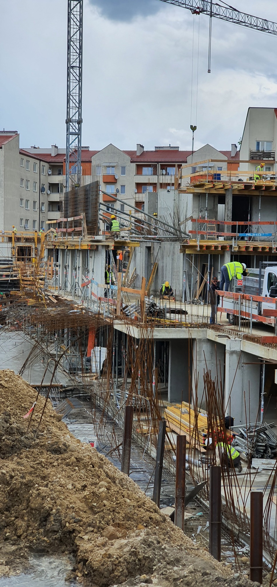 Wielopiętrowa konstrukcja żelbetowa w trakcie budowy, widoczne szalunki, zbrojenia i pracujący robotnicy w żółtych kamizelkach, w tle budynki mieszkalne i dźwig.
