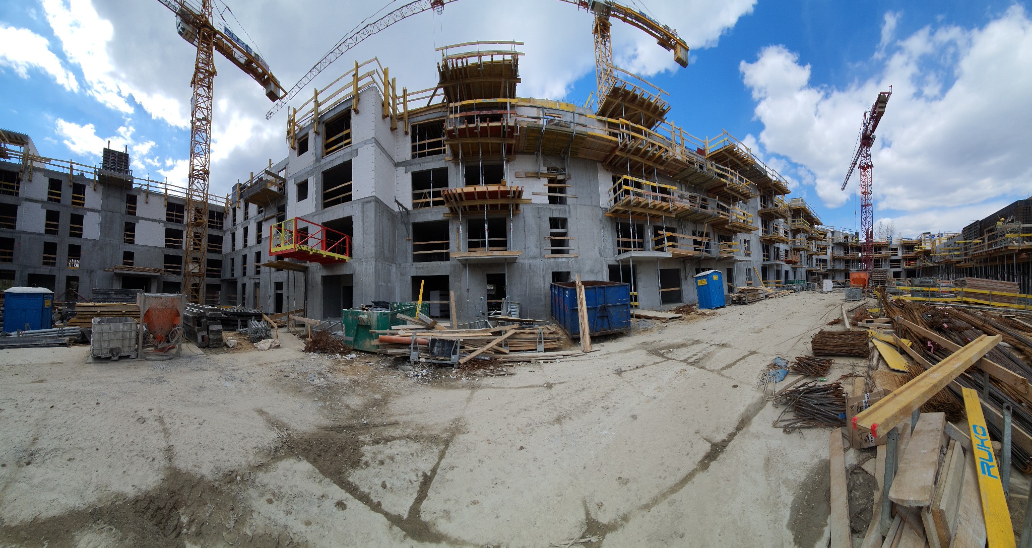 Panorama placu budowy wielopiętrowego budynku żelbetowego w trakcie wznoszenia, widoczne rusztowania, dźwigi i materiały budowlane.