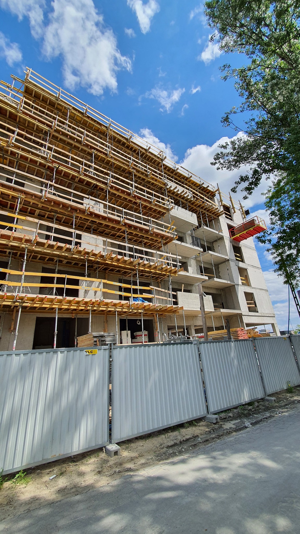 Wysoki budynek w trakcie budowy z widocznym szalunkiem drewnianym, metalowym ogrodzeniem i częściowo zachmurzonym niebem