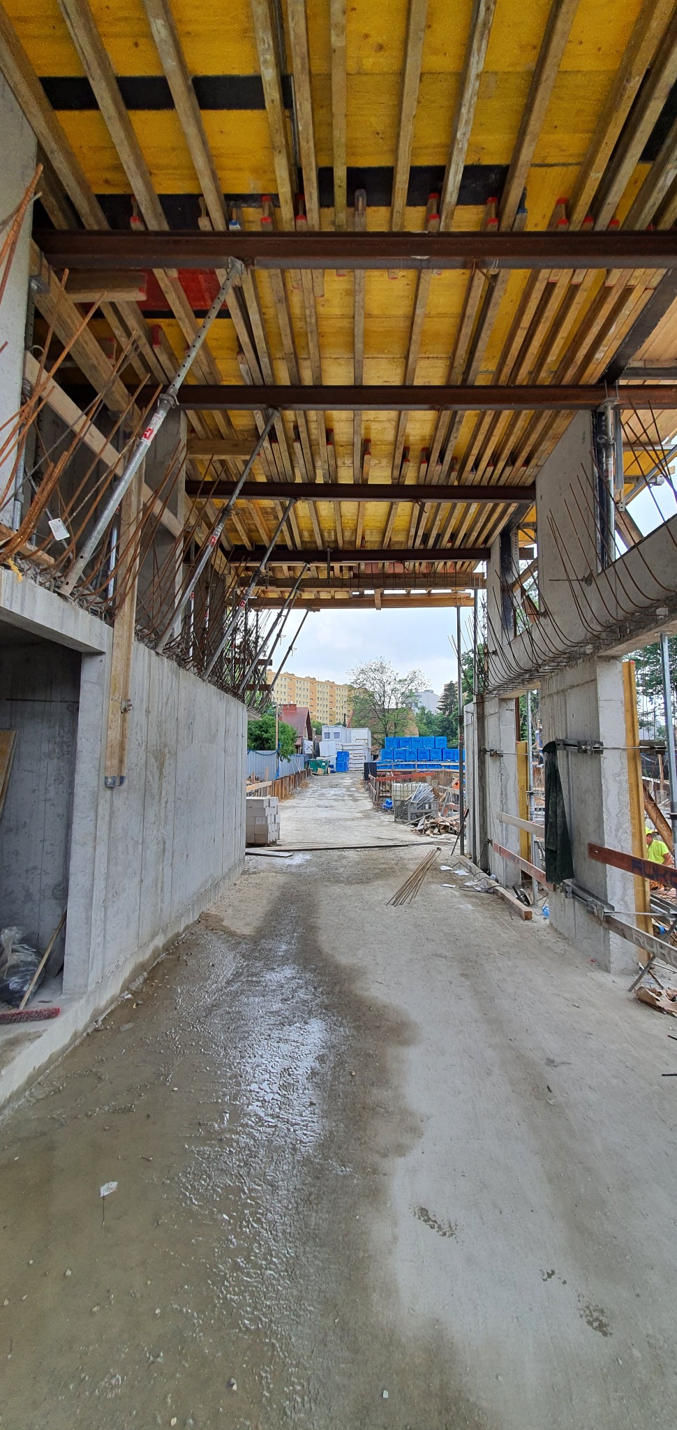 Wnętrze surowego budynku żelbetowego w trakcie budowy, widoczne szalunki stropowe z żółtymi płytami i stalowe belki, mokra posadzka betonowa, w tle budynki mieszkalne.