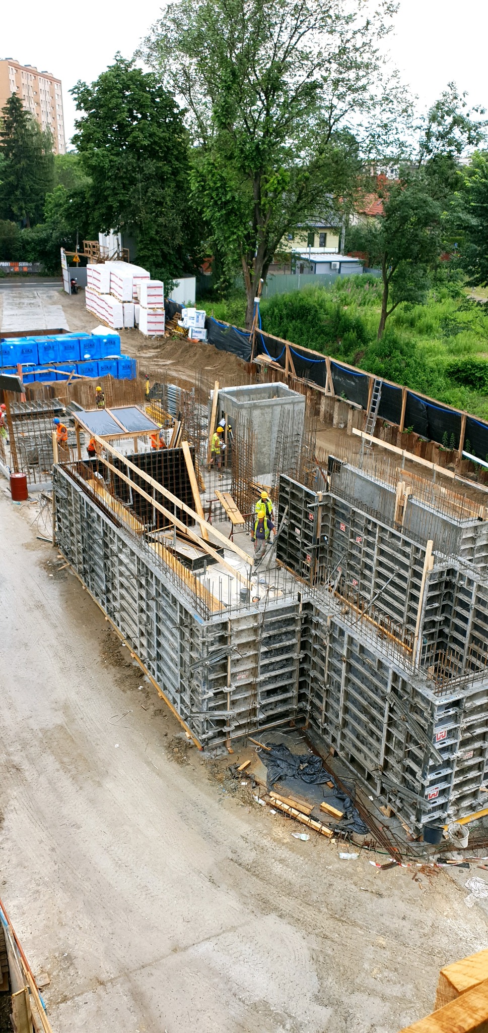 Wysoka perspektywa na plac budowy z widocznymi deskowaniami fundamentów, zbrojeniami i pracownikami w żółtych kamizelkach, w tle drzewa i budynki mieszkalne.