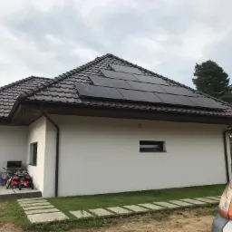 Dom jednorodzinny z brązowym dachem pokrytym panelami słonecznymi; widoczny fragment pomarańczowego samochodu dostawczego, zabawki dziecięce na tarasie i pochmurne niebo.