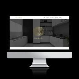 Monitor wyświetla stronę internetową z wizualizacją nowoczesnej kuchni w odcieniach szarości i bieli, z minimalistycznym logo firmy Luksann w złotym kolorze.