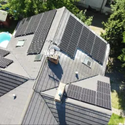 Widok z drona na dach domu z zainstalowanymi panelami słonecznymi; w tle basen, ogród i samochód.