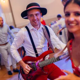 Uśmiechnięty basista w stroju góralskim, czarny kapelusz z czerwonym akcentem, biała koszula z szelkami, czerwone spodnie, grający na czerwonym basie podczas wesela, w tle tańczący goście.