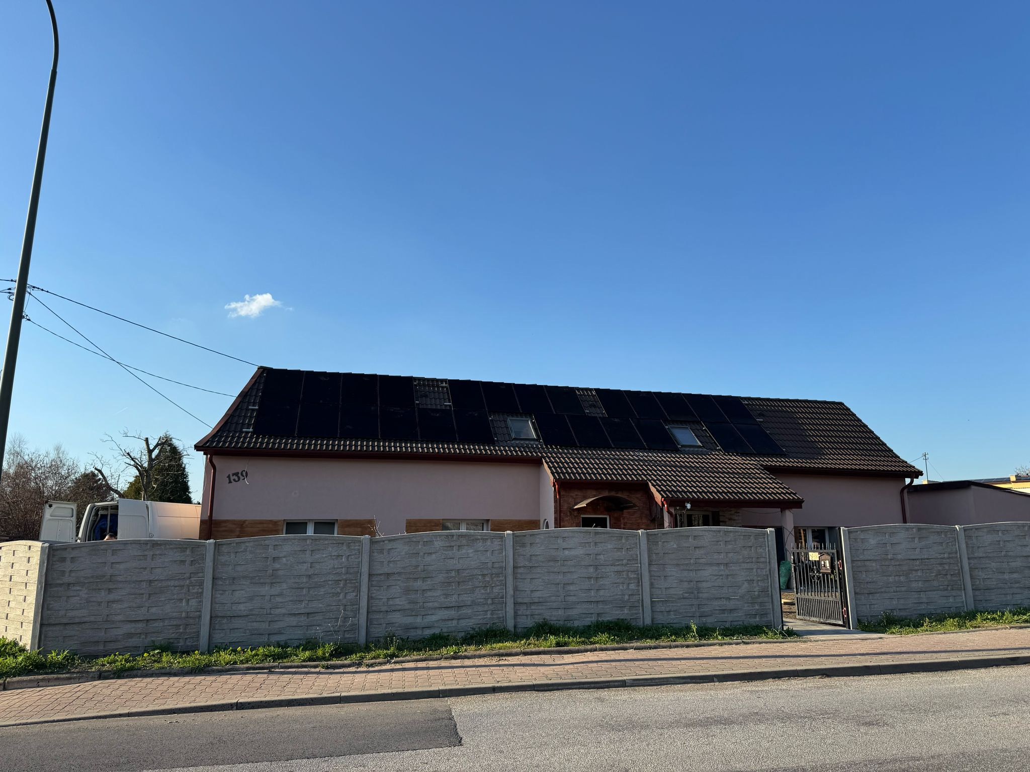Dom jednorodzinny z panelami słonecznymi na dachu, widoczny zza betonowego ogrodzenia. Numer domu 139, błękitne niebo w tle.