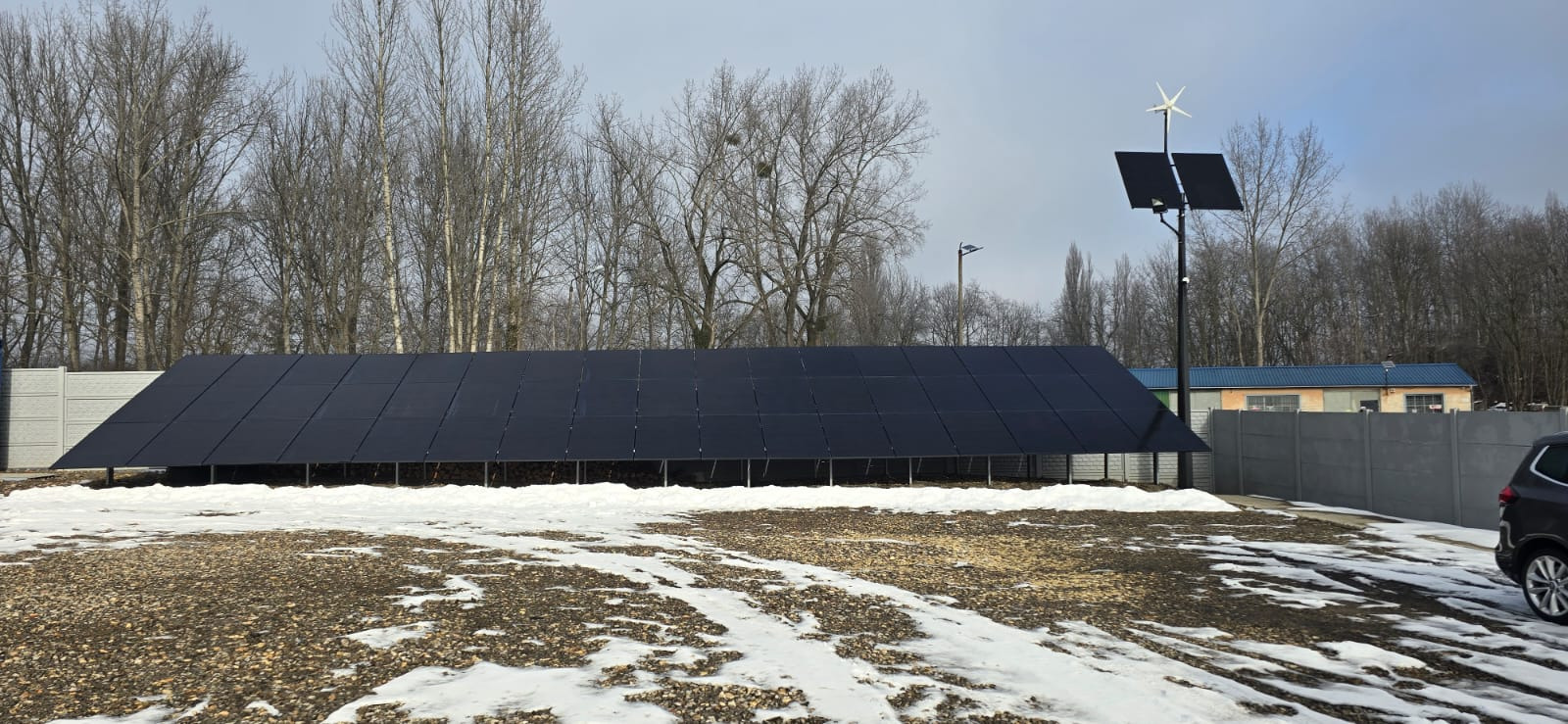 Rozległa instalacja fotowoltaiczna na gruncie, z dodatkowym panelem i turbiną wiatrową, otoczona drzewami i częściowo pokryta śniegiem.