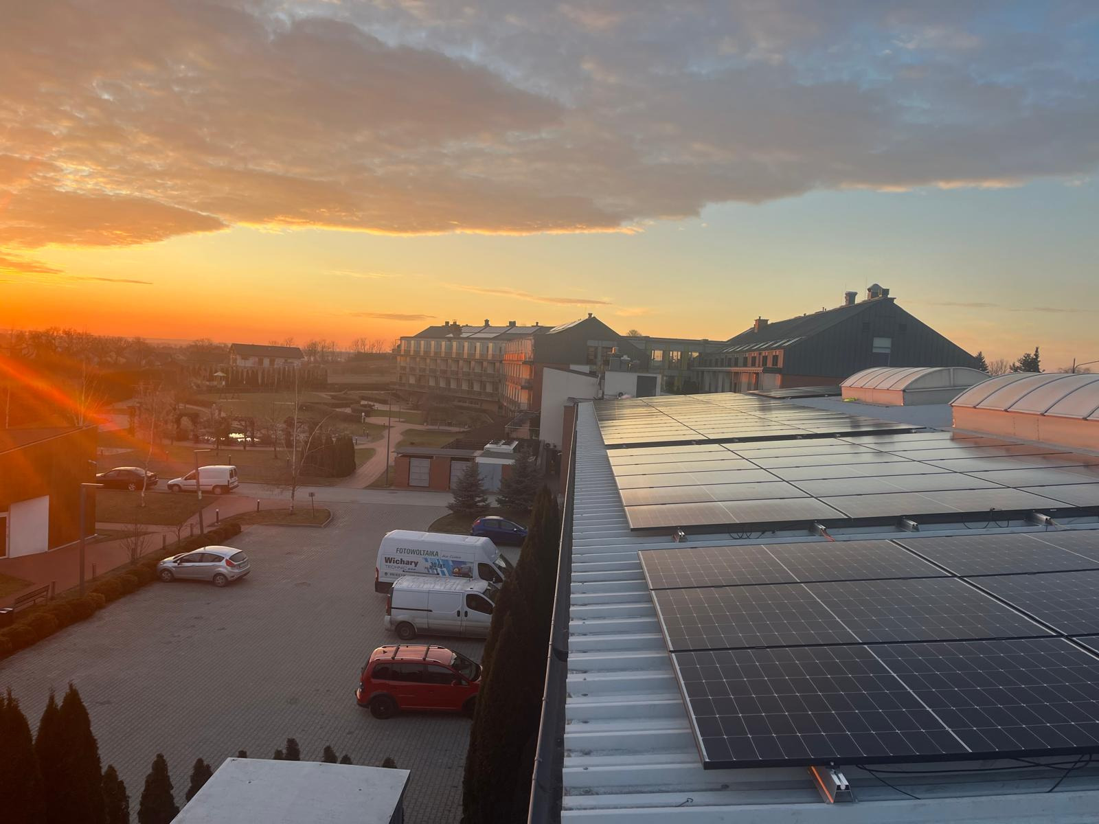 Panele słoneczne na dachu budynku oświetlone ciepłym, wschodzącym słońcem. W tle zabudowania i firmowe busy z logiem firmy w porannym krajobrazie.