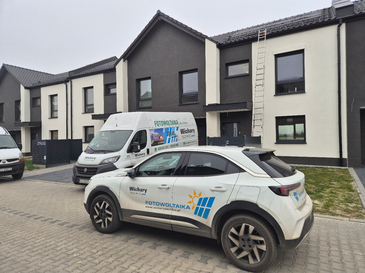 Biały samochód firmy z logo paneli słonecznych i van z napisem 'Fotowoltaika', zaparkowane przed nowoczesnymi domami z drabiną opartą o elewację.