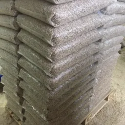 Stos worków z pelletem ułożonych na drewnianej palecie, widoczne drobne trociny na podłożu.