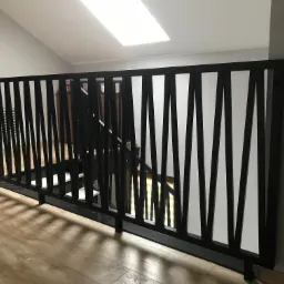 Nowoczesna, czarna, metalowa balustrada schodowa z pionowymi elementami o nieregularnych kształtach, zamontowana w jasnym wnętrzu z drewnianą podłogą i oknem dachowym.