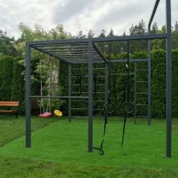 Metalowa konstrukcja placu zabaw z drabinką, drążkiem do podciągania i huśtawkami na tle zielonego trawnika i żywopłotu.