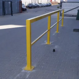Ż&oacute;łta, metalowa barierka ochronna przy rampie załadunkowej, zamontowana na kostce brukowej, z widocznym śmietnikiem i bramą w tle.