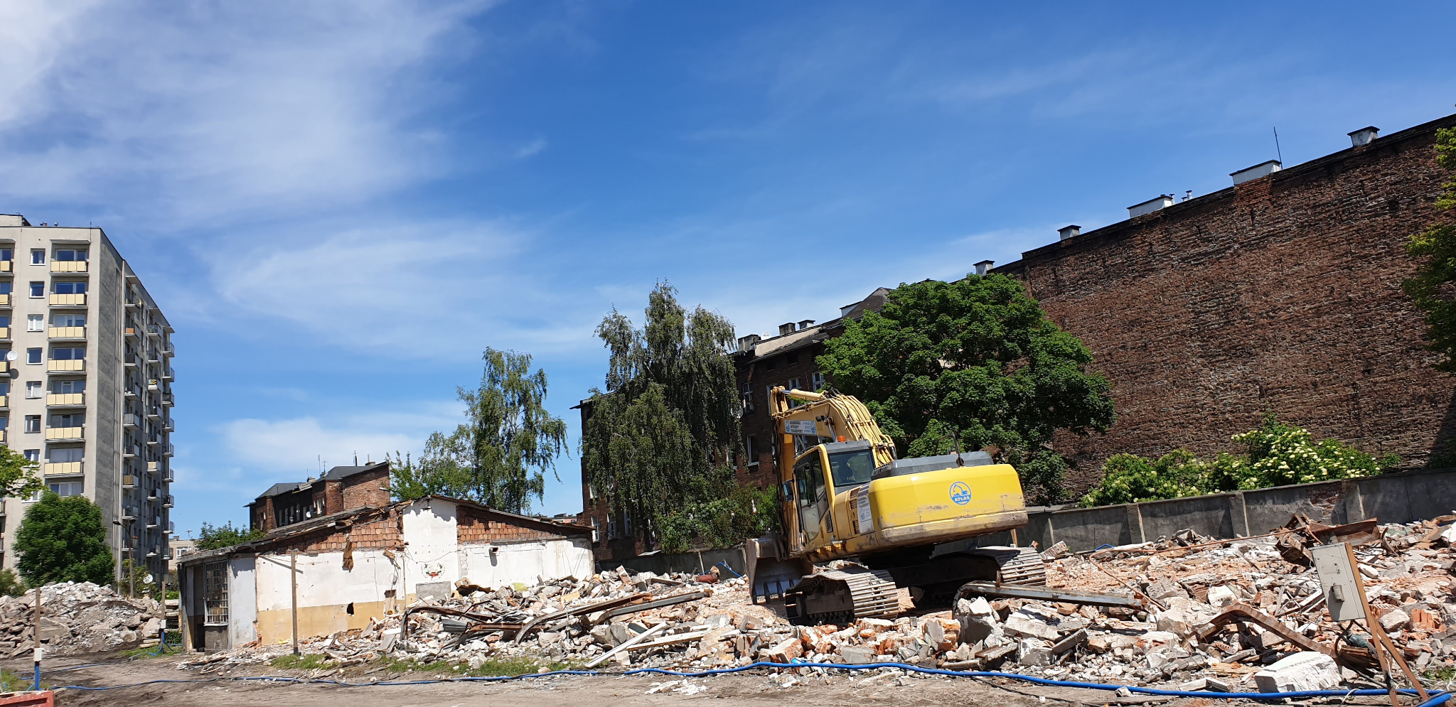 Żółta koparka gąsienicowa na gruzowisku po rozbiórce budynku, z widocznym blokiem mieszkalnym w tle i fragmentem wysokiego ceglanego muru.