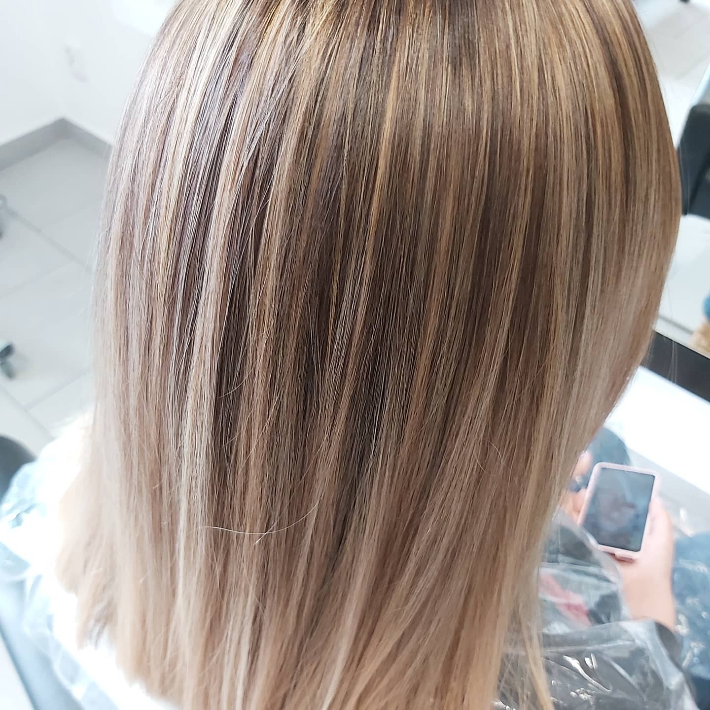 Blond włosy z delikatnymi refleksami, widoczne od tyłu, klientka trzyma telefon w dłoni.