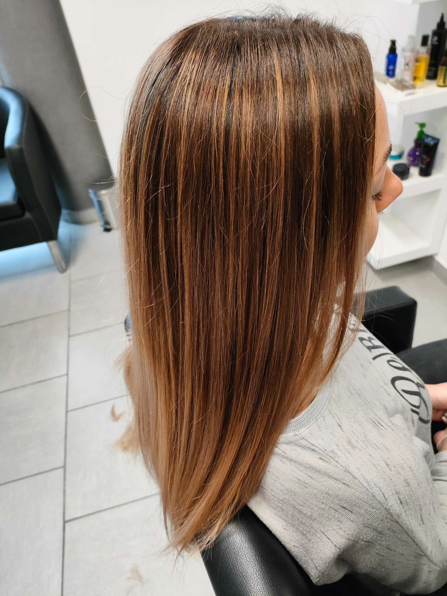 Długie, proste włosy z brązowymi refleksami, widoczne z tyłu głowy klientki w salonie, z delikatnym gradientem koloru ku końcówkom.