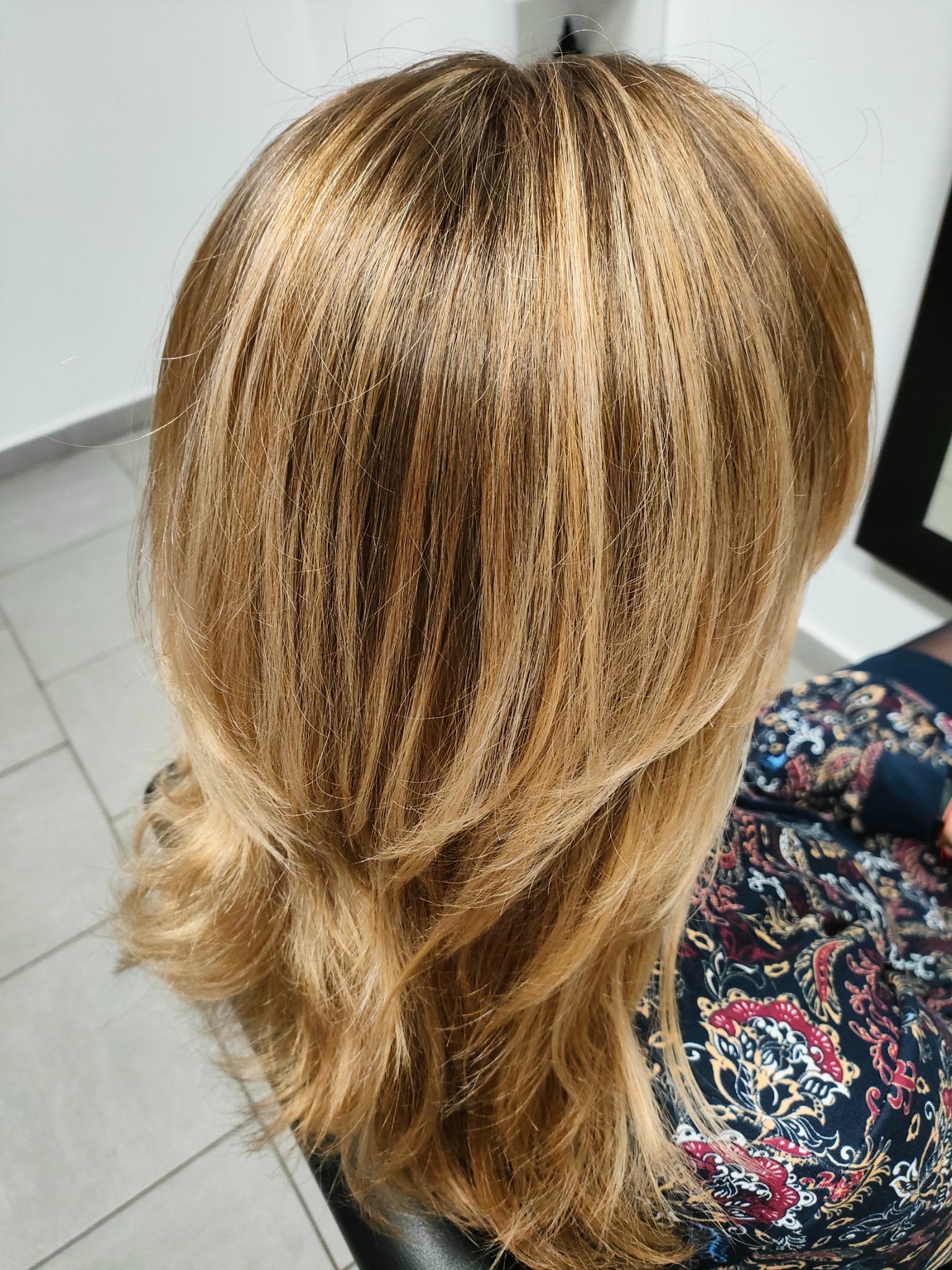 Tył głowy kobiety z blond włosami z pasemkami, delikatnie pocieniowane, widoczny wzorzysty materiał na ubraniu.