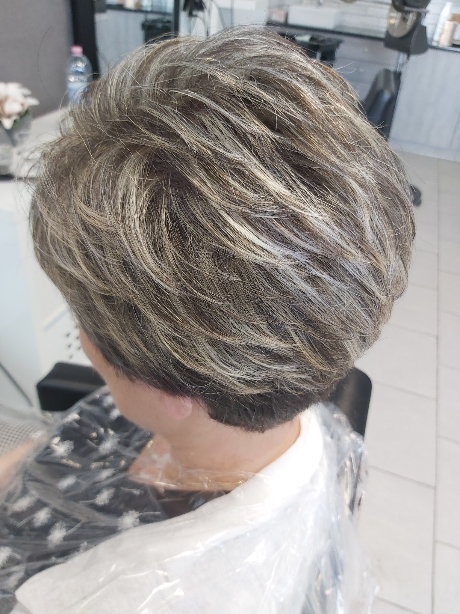 Krótkie, warstwowe cięcie włosów w odcieniach szarości i blondu, widoczne od tyłu, z plastikową peleryną ochronną na ramionach.