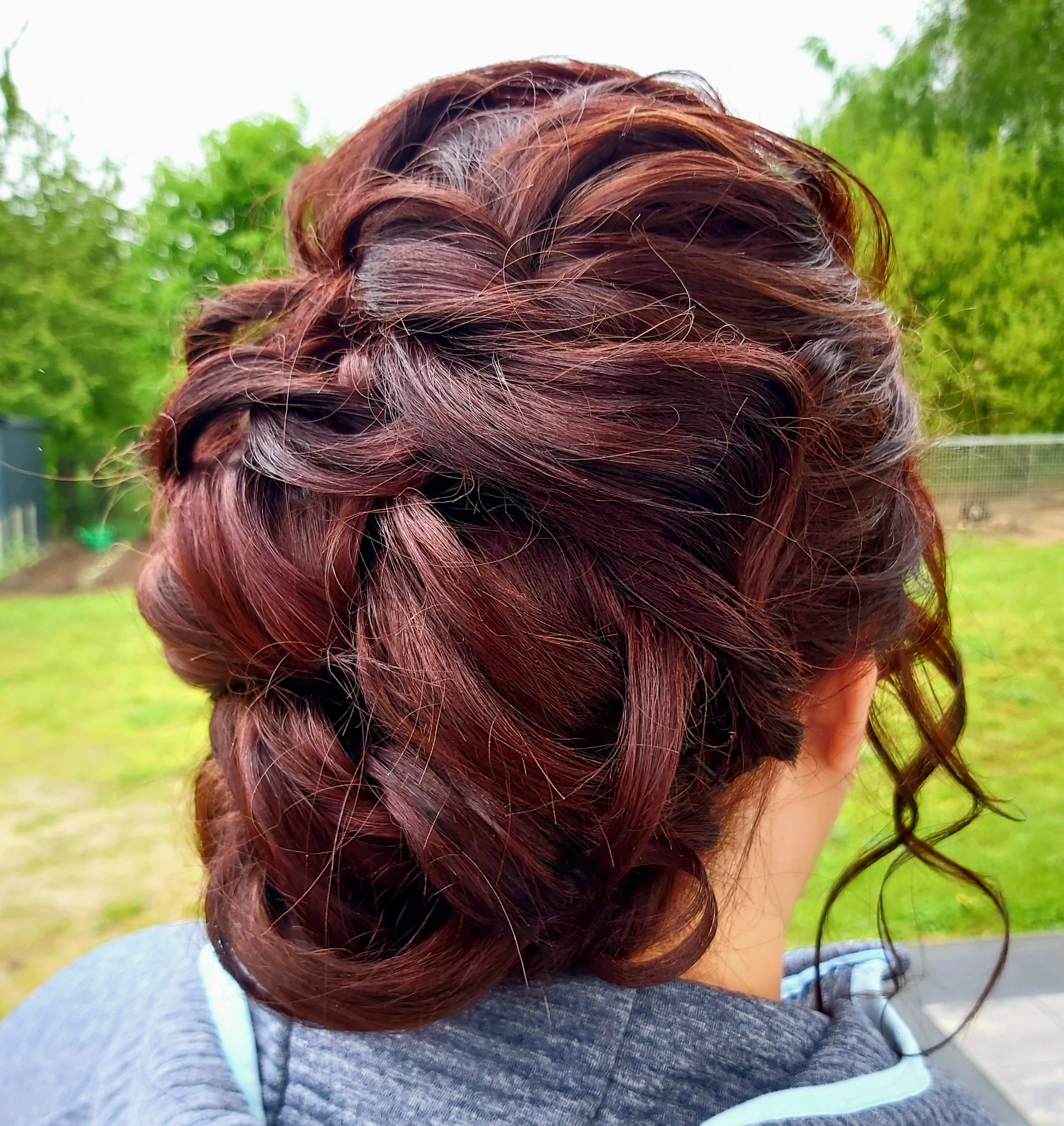 Upięcie z ciemnych, kręconych włosów z widocznymi warkoczami, na tle zieleni.