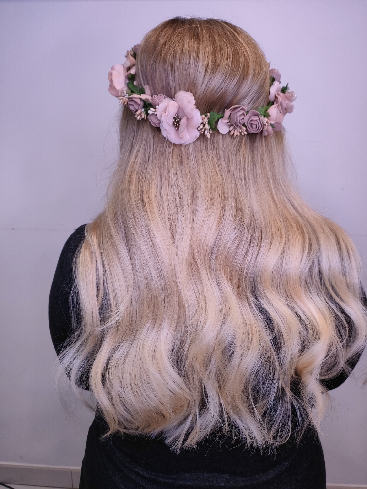 Długie, falowane, blond włosy z tyłu głowy, ozdobione delikatnym, różowym wiankiem z kwiatów.