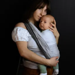 Kobieta z długimi włosami trzyma niemowlę zawinięte w szaro-biały materiał, na ciemnym tle.