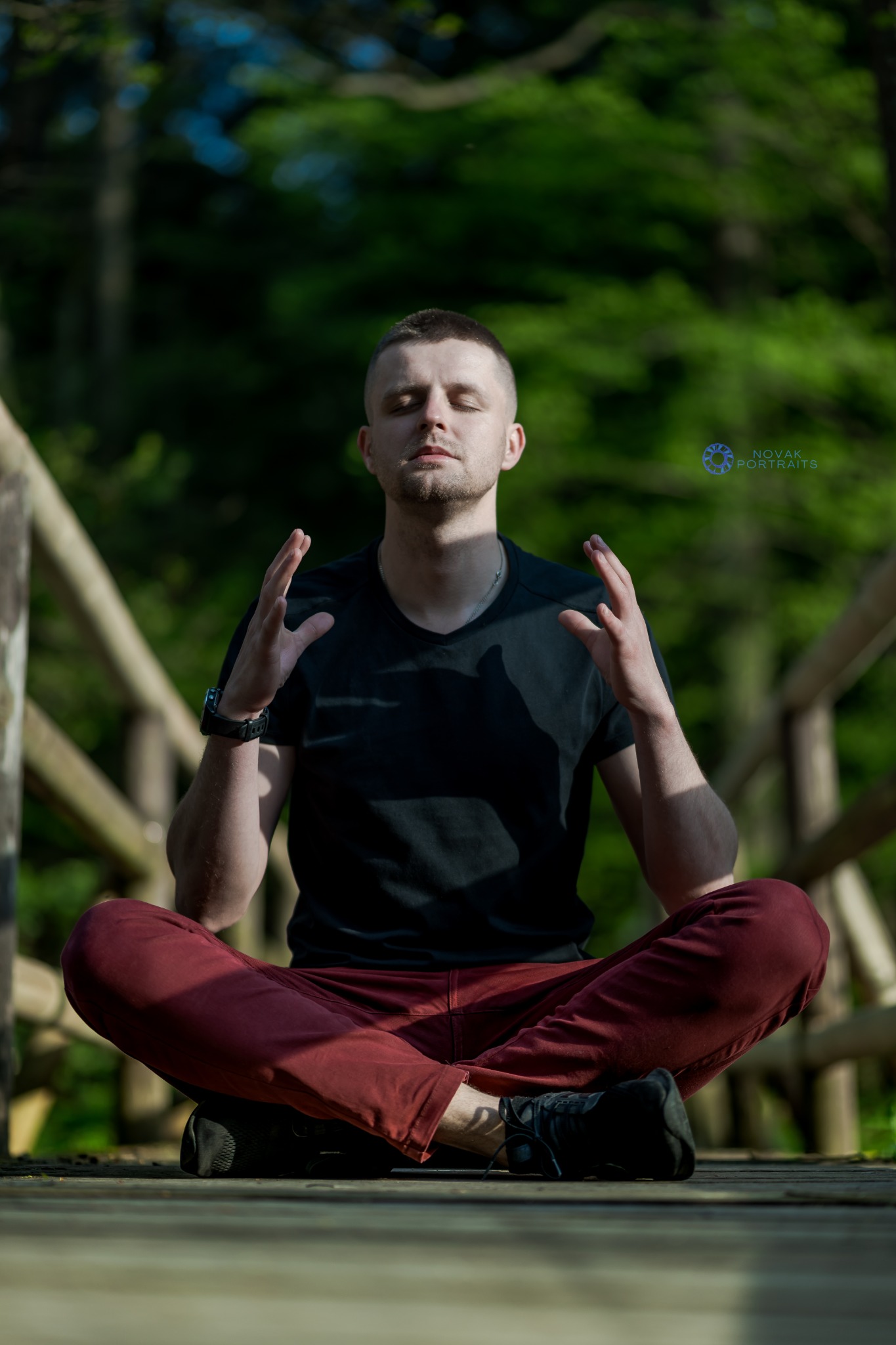 Mężczyzna w czarnej koszulce i bordowych spodniach siedzi w pozycji lotosu na drewnianym moście, z rękami uniesionymi w geście medytacji, z zaciśniętymi powiekami, w tle zieleń drzew.