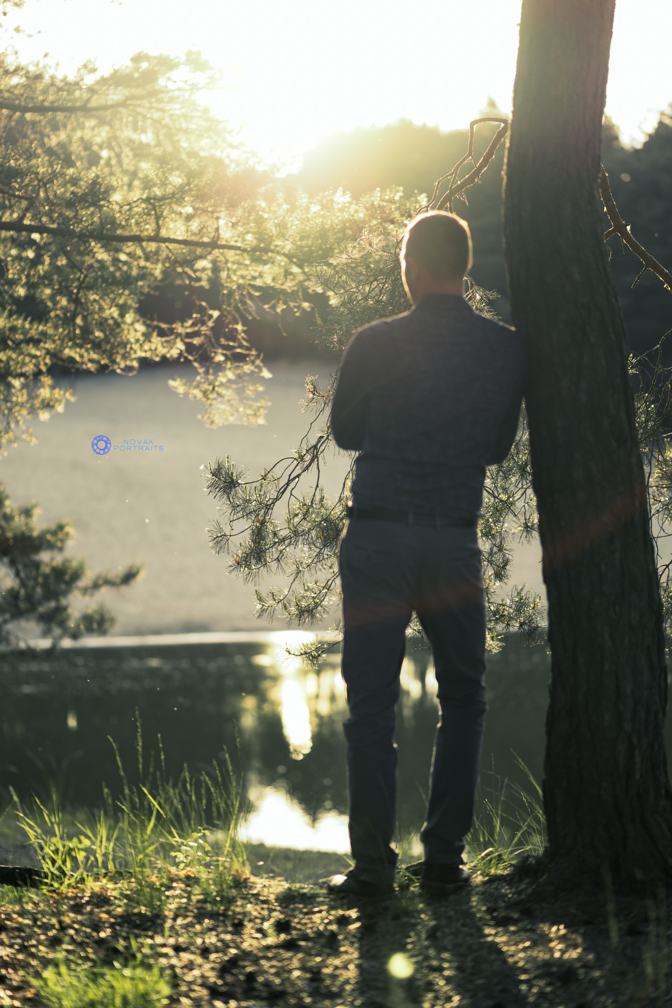 Mężczyzna oparty o drzewo nad jeziorem w promieniach zachodzącego słońca, widok z tyłu, sylwetka na tle błyszczącej wody.