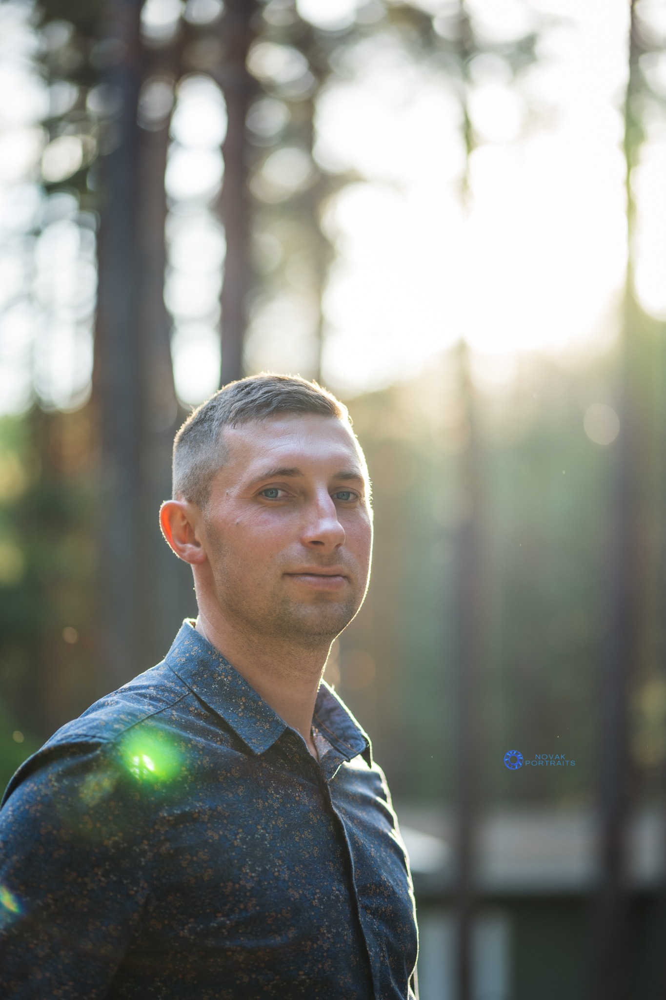 Portret mężczyzny w niebieskiej koszuli w drobny wzór, na tle rozświetlonego słońcem lasu, delikatne bokeh w tle.