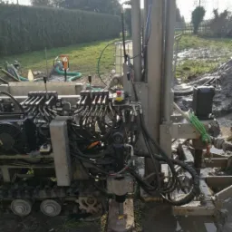 Wiertnica na gąsienicach, przygotowana do wiercenia, z widocznymi przewodami hydraulicznymi i elementami sterującymi, otoczona błotnistym terenem z trawą i żywopłotem w tle.