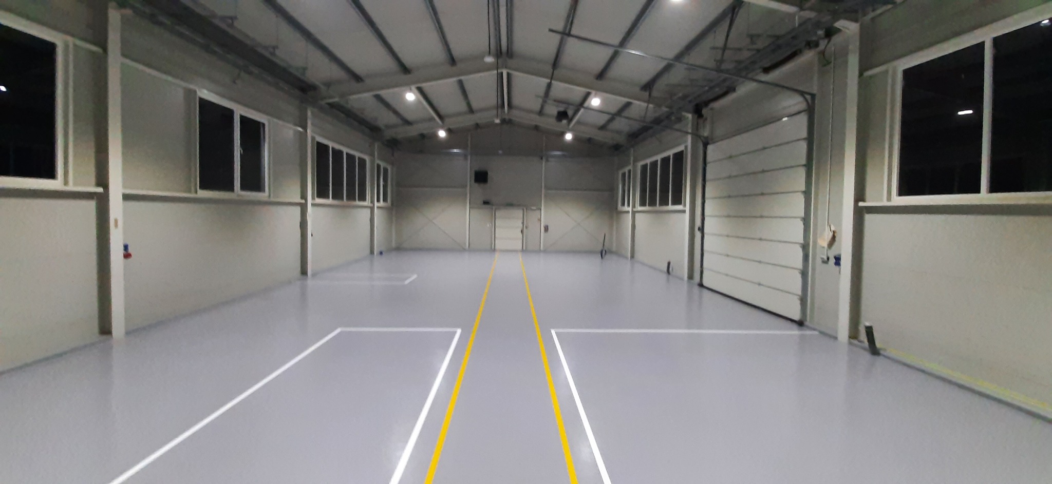 Jasnoszara, gładka posadzka w hali przemysłowej z wyznaczonymi żółtymi i białymi liniami, widok na bramę garażową i okna.