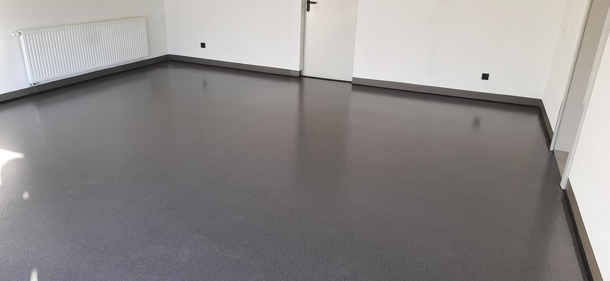 Gładka, ciemnoszara powierzchnia podłogi w jasnym pomieszczeniu z białymi ścianami, grzejnikiem i drzwiami. Widoczny ciemny cokół przypodłogowy.