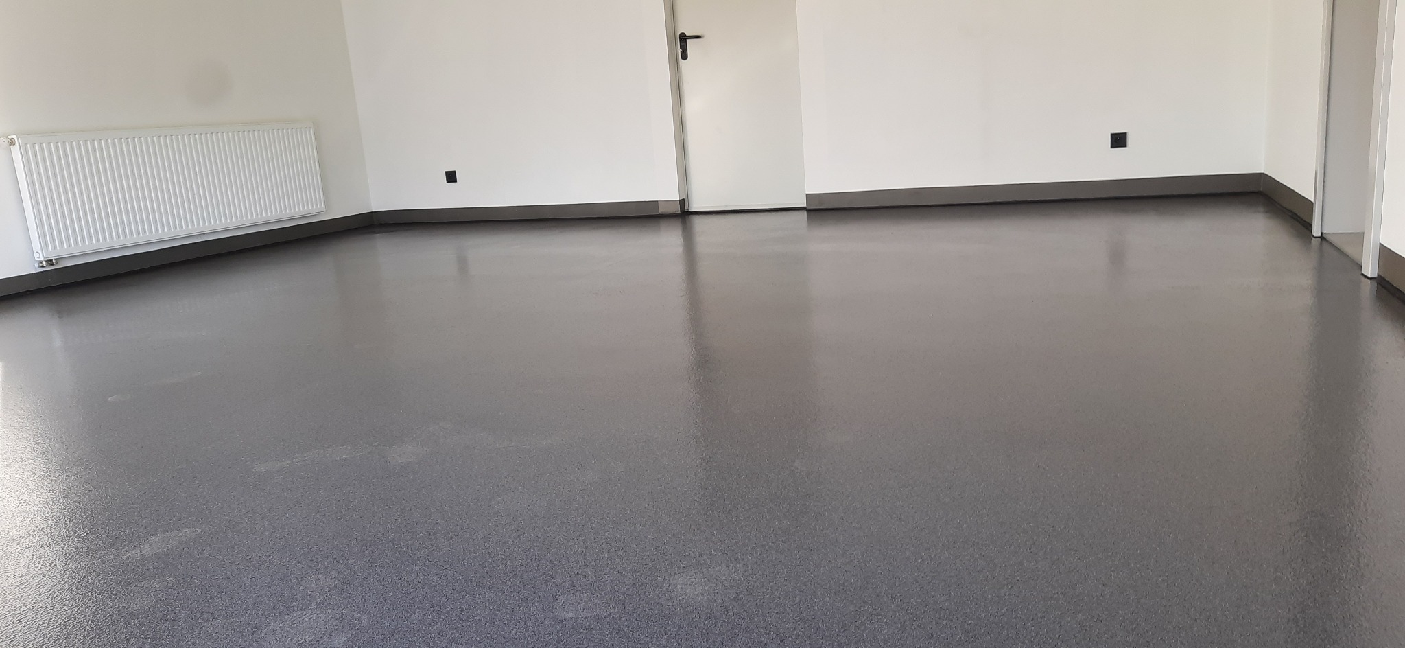 Gładka, szara posadzka w jasnym pomieszczeniu z białymi ścianami, grzejnikiem i drzwiami.