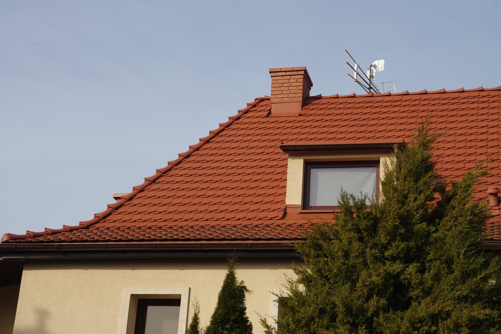 Czerwony dach z dachówki karpiówki, komin z cegły, antena satelitarna na dachu, widok z dołu na tle jasnego nieba, iglaki w ogrodzie.