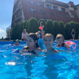 Grupa dzieci w basenie na tle budynku z czerwonym dachem i zielonymi tujami, jedno dziecko z psem i opiekunem stoi obok basenu.